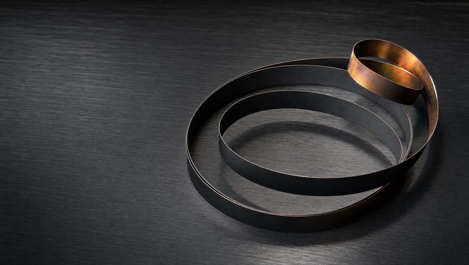 PTFE coated endless steel bands coiled loosely, showing the matte black coated face against the bronze back left by the curing oven.